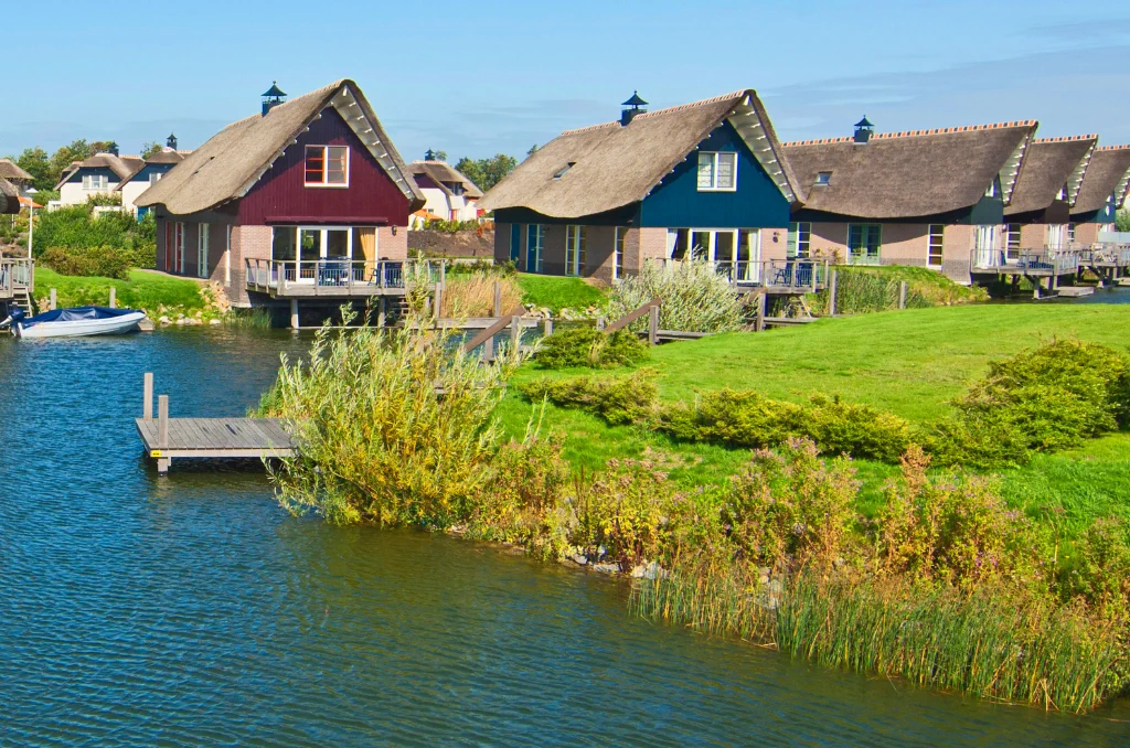 Eine Reihe von bunten Häusern mit Reetdächern am Ufer eines ruhigen Kanals an einem sonnigen Tag.
